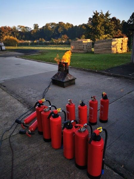 Mehrere rote Feuerlöscher stehen aufgereiht auf einem Übungsgelände im Freien. Im Hintergrund brennt eine kontrollierte Flamme auf einer Trainingsvorrichtung zur Feuerlöscher-Ausbildung.