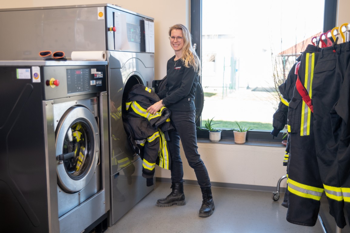 Eine Frau lädt Feuerwehrschutzkleidung in eine Industriemaschine in einem hellen Waschraum mit großen Fenstern.