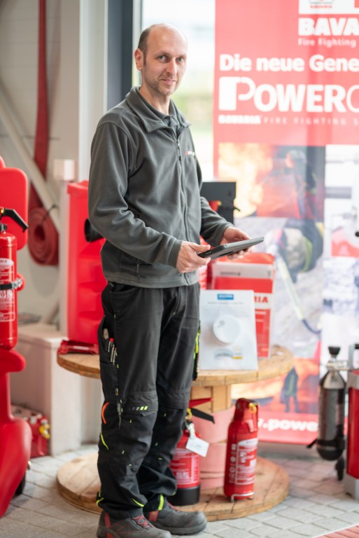 Ein Mann in Arbeitskleidung hält ein Tablet in einem Showroom für Brandschutztechnik mit Feuerlöschern und Werbebannern im Hintergrund.