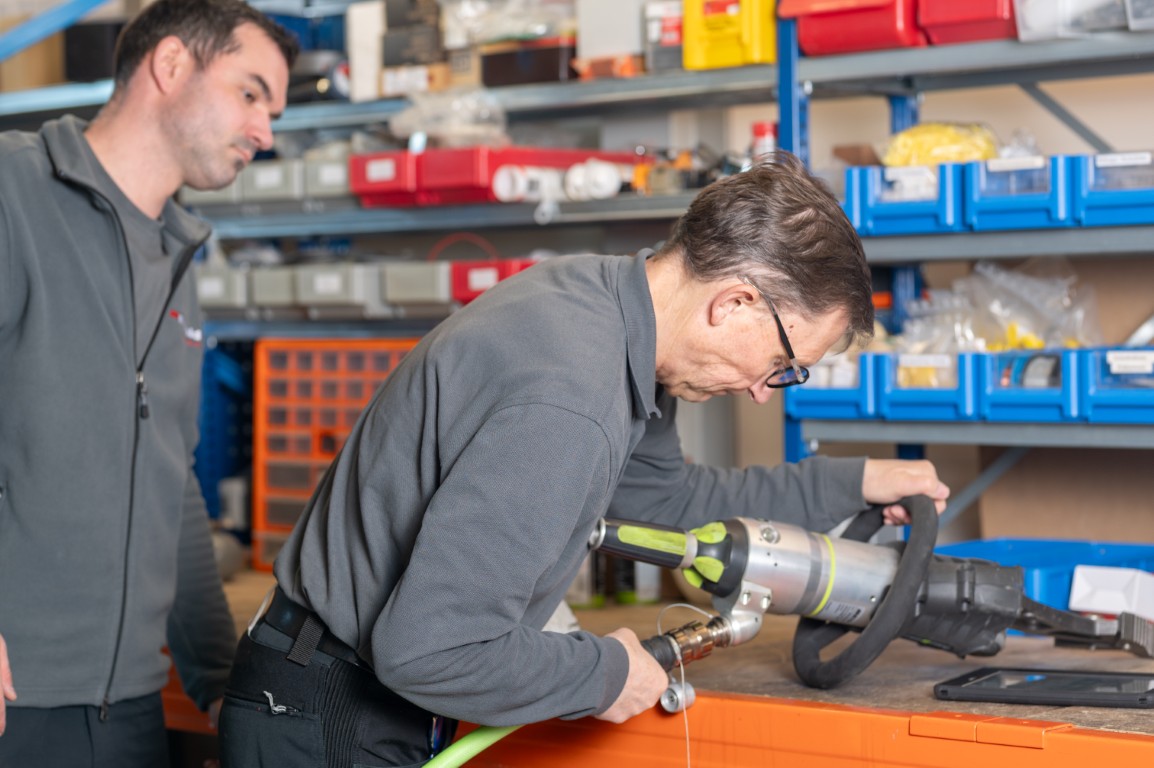 Zwei Männer arbeiten in einer Werkstatt – einer prüft ein hydraulisches Rettungsgerät, der andere beobachtet. Im Hintergrund Regale mit Ersatzteilen und Werkzeugen.